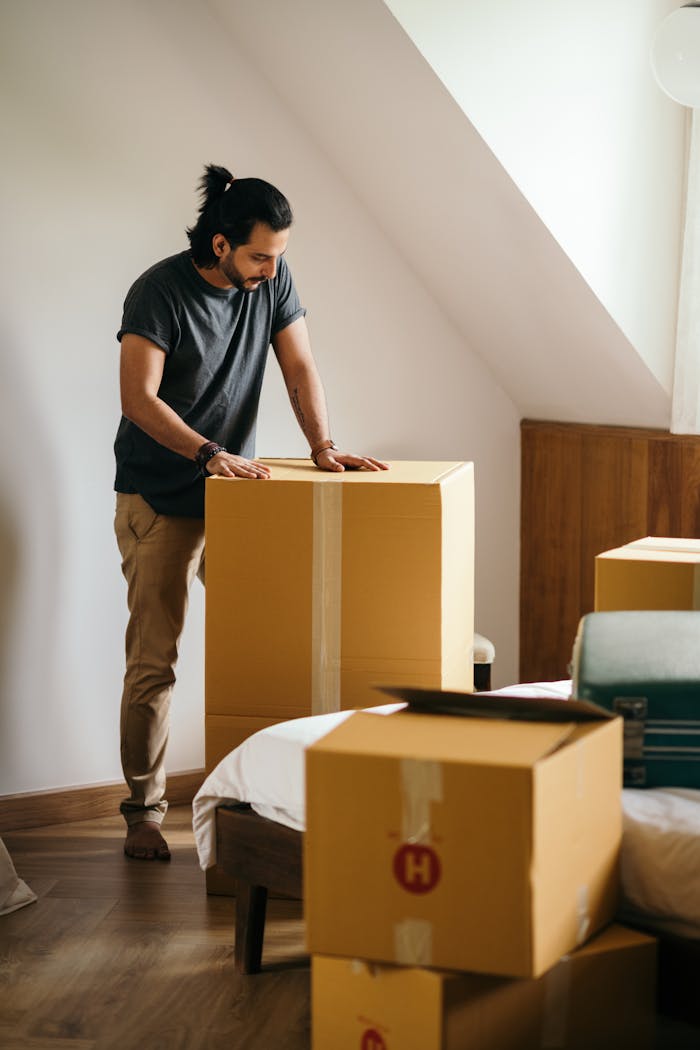 about-img Ethnic man packing belongings in cardboard boxes while standing in modern apartment with white wall and looking down while preparing for relocation